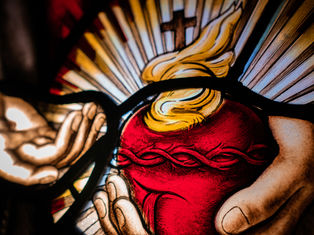 Stained glass art depicting hands holding a red heart with a crown of thorns, flames, and rays in the background, creating a luminous effect.