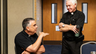 Two deacons in black clerical shirts talk in a meeting room, one seated gesturing, the other standing with a water bottle.