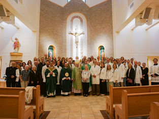 A group of people, including clergy in green robes and individuals in white coats, pose in a church with a crucifix and statues in the background.