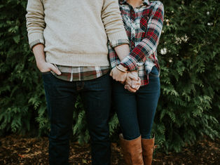 A couple holds hands, wearing casual fall attire with plaid and jeans. They're in front of green foliage, setting a cozy, intimate mood.