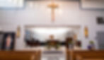 Church interior with altar, cross and religious statues. Bright lighting, floral decorations, wooden pews, and a serene atmosphere.