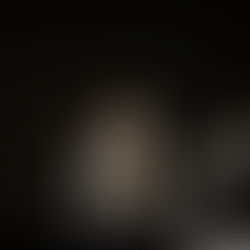 A clergyman in ornate robes speaks at a podium on a dimly lit stage, with seated attendees in white robes. Candle visible, somber mood.