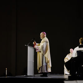 A clergyman in ornate robes speaks at a podium on a dimly lit stage, with seated attendees in white robes. Candle visible, somber mood.
