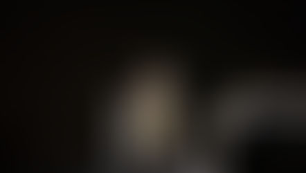 A clergyman in ornate robes speaks at a podium on a dimly lit stage, with seated attendees in white robes. Candle visible, somber mood.