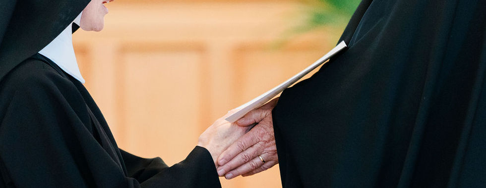 A religious sister kneels before her superior during the profession of vows ceremony as her hands are gently received in prayer.