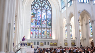 Priest in purple robe speaks from pulpit to seated congregation in bright cathedral with tall stained-glass windows