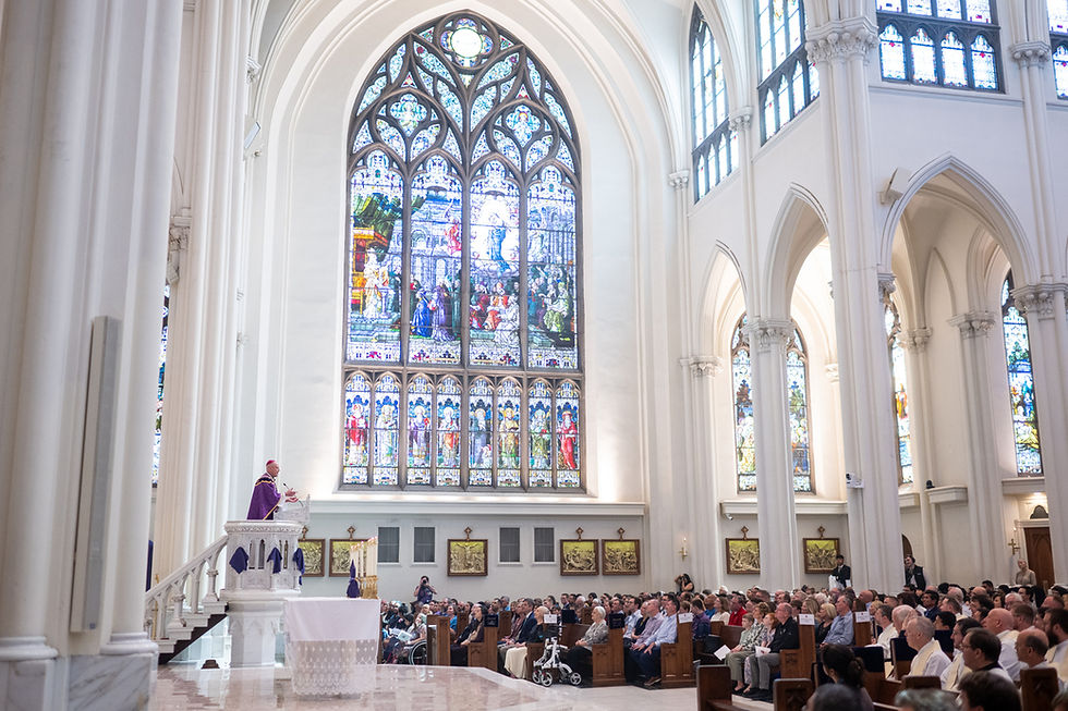 Priest in purple robe speaks from pulpit to seated congregation in bright cathedral with tall stained-glass windows