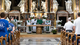 Catholic Mass in an ornate church, priests in green vestments at the altar while the congregation stands in pews before marble statues.