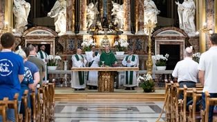 Catholic Mass in an ornate church, priests in green vestments at the altar while the congregation stands in pews before marble statues.