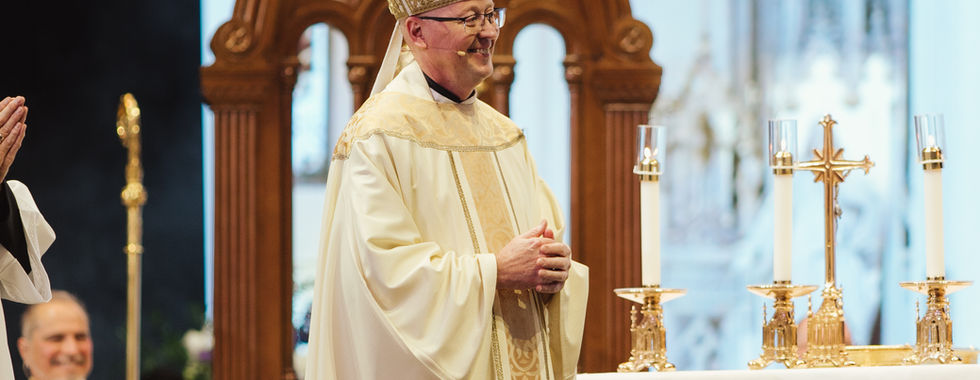 A bishop in elaborate robes stands at an altar, smiling during a ceremony. The setting is ornate with gold and floral accents.