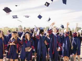 Graduates in blue gowns toss caps at an outdoor ceremony, smiling under a bright sky.