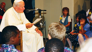 A person in white robes speaks into a mic, holding a paper. Children in colorful outfits listen attentively in a room with chairs.
