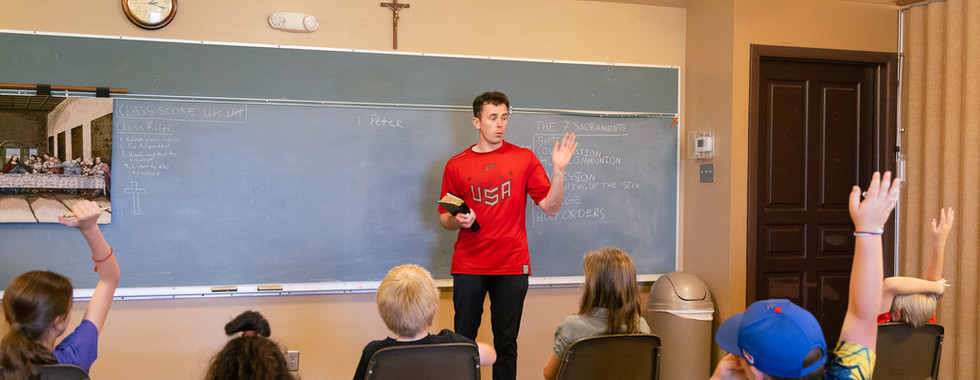 Teacher in red shirt holds a book, standing by a chalkboard with class rules. Kids raise hands. Classroom setting with religious decor.