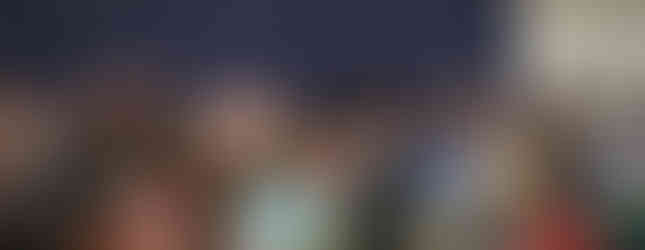 People seated at tables in a crowded hall, having conversations. The backdrop is blue, and the mood is lively. Water bottles on tables.