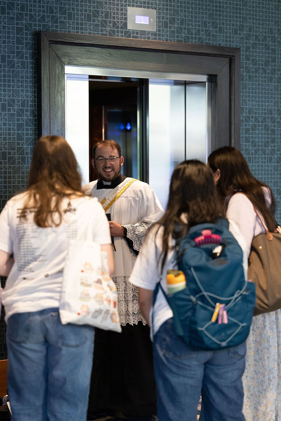 Smiling priest in white robe greets three women with backpacks in a patterned elevator lobby.