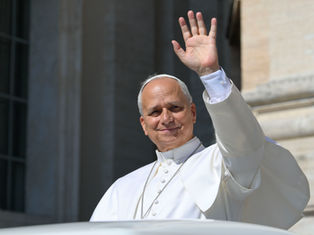El papa León, con sotana blanca, saluda con la mano desde un balcón; fondo de piedra y luz soleada.
