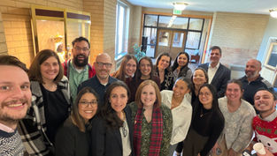 Smiling group selfie of coworkers in a brick hallway near an EXIT sign, posing closely in a bright indoor lobby