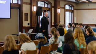A speaker addresses an attentive audience in a wood-paneled room. A laptop and projector screen are visible, creating an academic atmosphere.