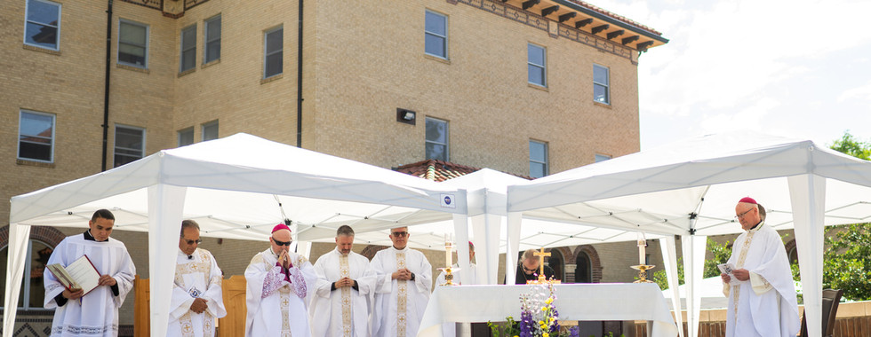 Outdoor Catholic Mass with priests in white vestments under canopies beside a flower-decorated altar and brick building.