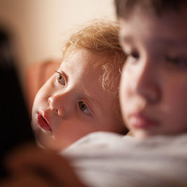 Two young children in warm indoor light, one blond child gazing upward while the other sits blurred in the foreground.