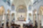 Crowded cathedral service with congregation standing in a bright Gothic church, stained glass, white arches, and purple altar drapes