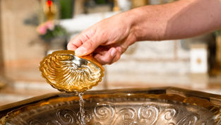 Hand pours water from an ornate gold shell cup into a basin in a softly lit interior, with warm reflective tones.