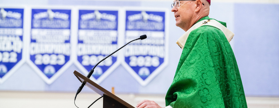 Bishop in green vestments speaks at a podium in a church gym, with blue banners reading 2022 and 2026 behind him.