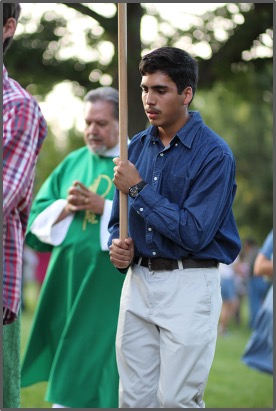 Young man in blue shirt holds a pole in a park, leading a procession. A man in green robes follows, suggesting a religious event.
