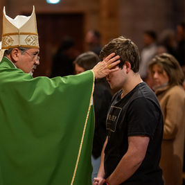 A bishop in green robes blesses a young man in a church, touching his forehead. The congregation is visible in the background.