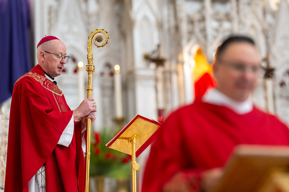 A bishop in red robes holds a golden staff in a church, with ornate white background. Another clergy in red is blurred in the foreground.