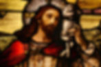Stained glass depicts a bearded man with a halo holding a lamb, wearing red and white robes. The scene is serene with a rural backdrop.