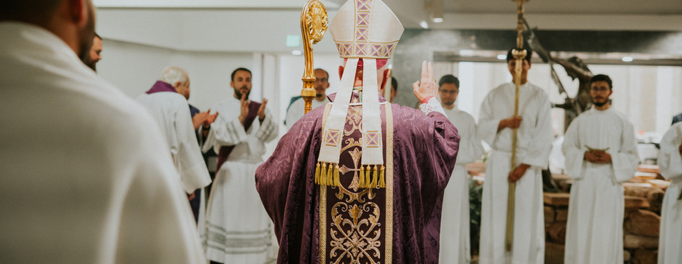A bishop in ornate purple robes performs a blessing ceremony. Clergy in white robes stand in a circle inside a church, creating a solemn mood.