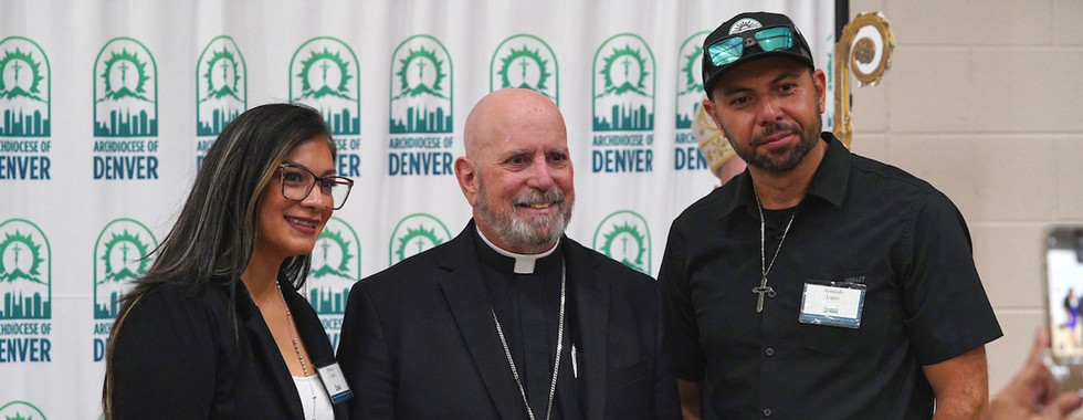 Three people pose smiling in front of an "Archdiocese of Denver" backdrop. The setting is bright and formal with a cheerful mood.