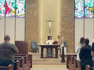 Priest at altar in church with a crucifix above, leading a service. Congregation stands in pews. Stained glass windows and brick wall backdrop.