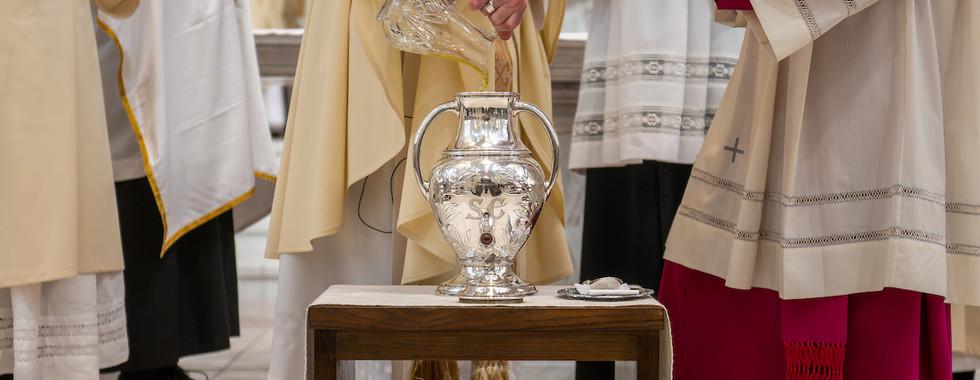Clergy in cream robes pour liquid into a silver urn labeled SC. One holds a red book. Background shows more robes in a church setting.