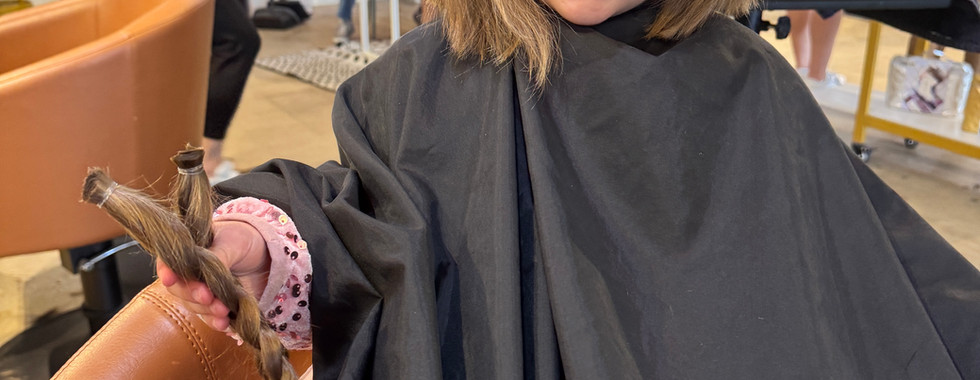 Young girl in a salon chair smiles while holding a freshly cut braid of her hair.