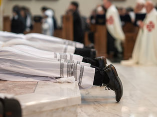 Men in white robes lie prostrate on a marble floor during a religious ceremony, surrounded by blurred figures in formal attire.