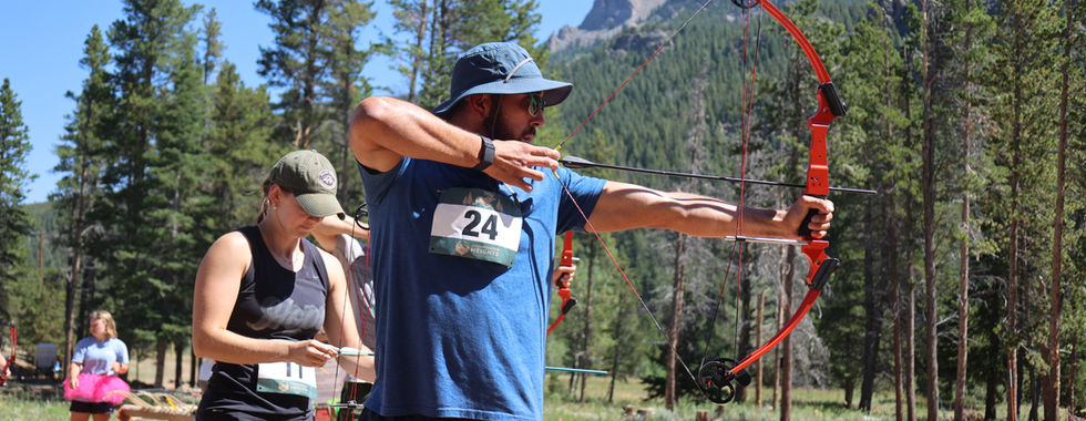 Man in blue hat aims a red recurve bow at an outdoor archery range, with a woman behind him and pine mountains under a clear sky.