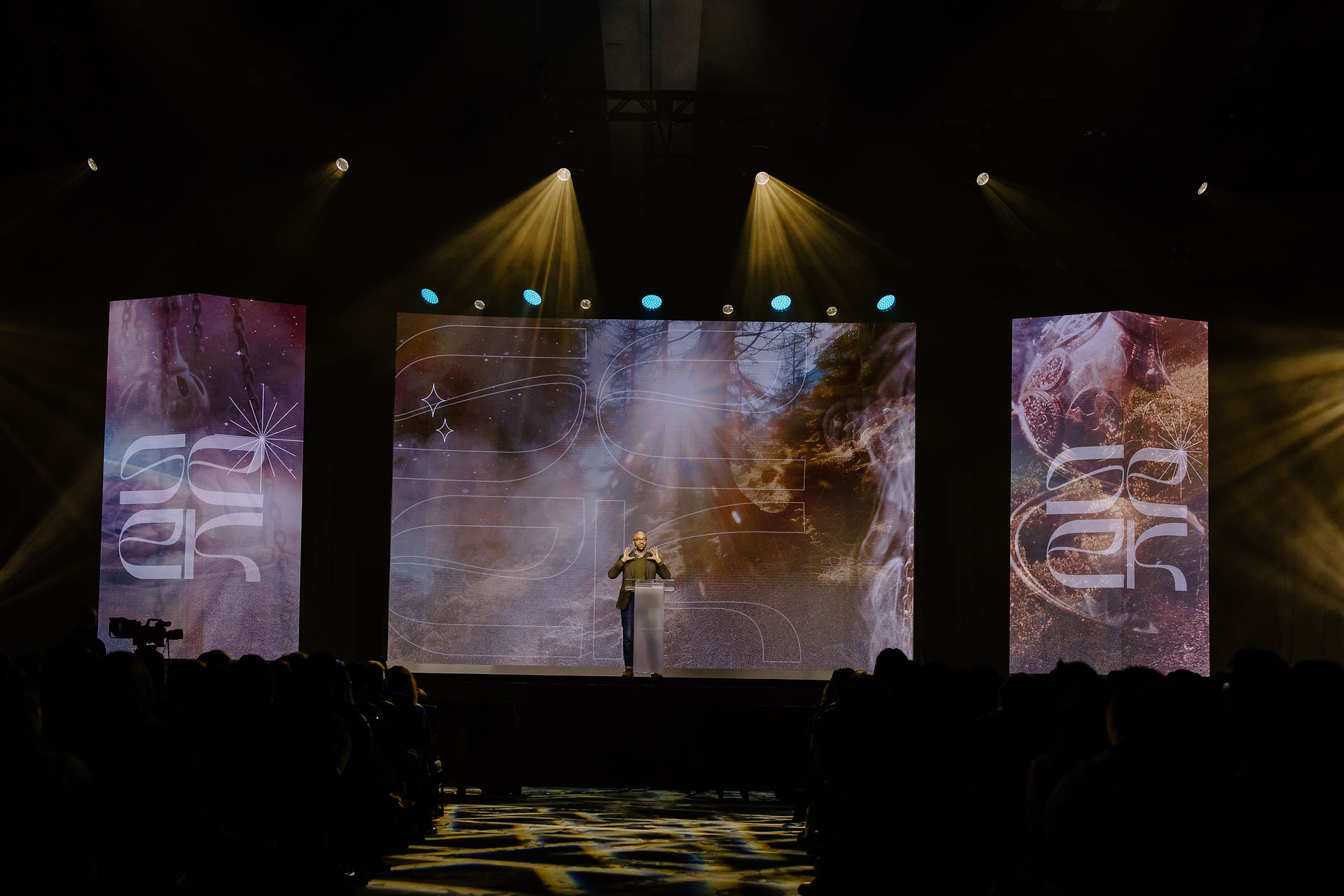 Speaker on stage with a podium, flanked by large colorful screens displaying abstract designs. Audience in shadow, illuminated by spotlights.
