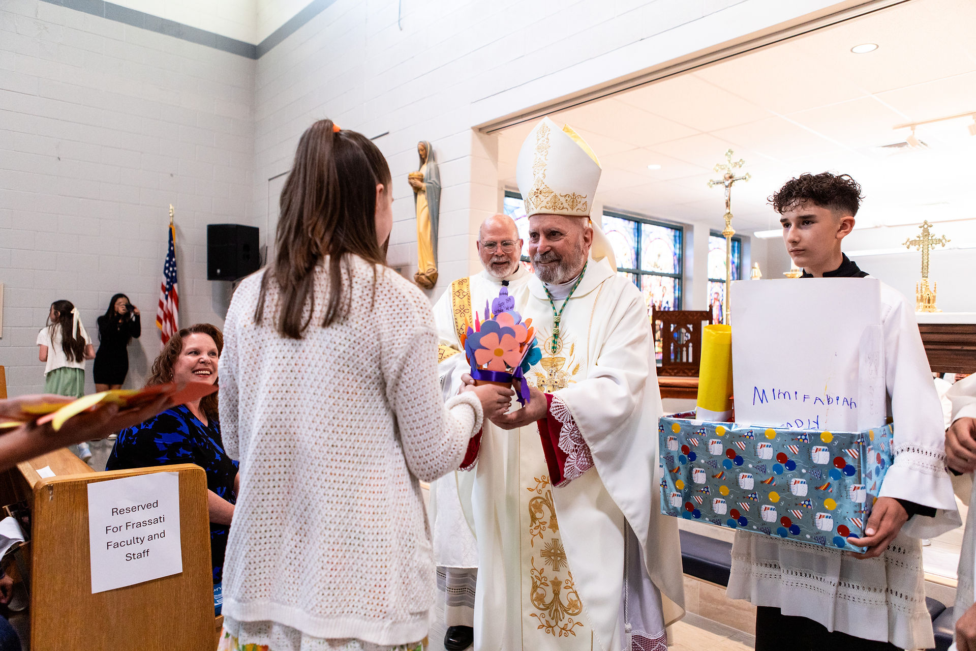 A bishop in white robe accepts colorful paper flowers from a girl in a church. Nearby, a boy holds a gift box. Reserved seating sign visible.
