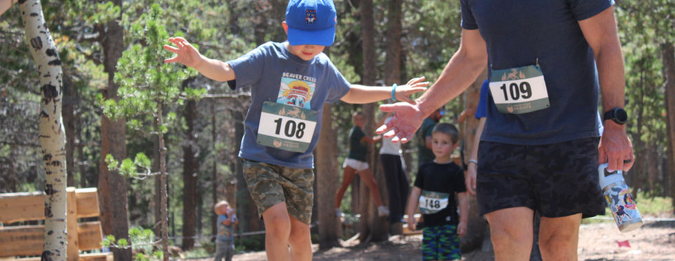 Boy balancing on a log in a forest race, reaching for an adult’s hand; bibs 108 and 109, smiling outdoors.