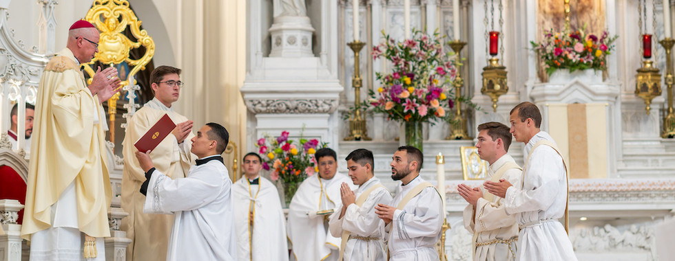 Priests in white robes kneel in a church, facing a bishop in cream robes beside an altar adorned with flowers and candles. Gothic architecture.