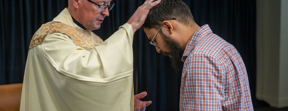 A clergy member in a robe blesses a man in a plaid shirt, pressing his hand on the man's head. Dark curtain backdrop.