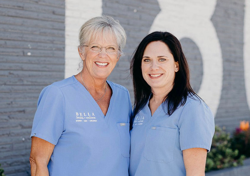 Two smiling women in blue scrubs with "BELLA health + wellness" text. They stand against a gray brick wall with white lettering.
