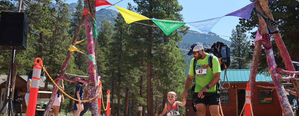 Man with backpack and small child run hand in hand under colorful bunting at a forest trail race finish, mountains behind