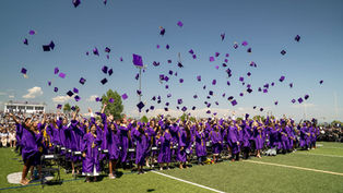 Graduates in purple gowns celebrate by tossing caps into the air on a sunny day at a stadium. Crowd watches; mood is joyful and festive.