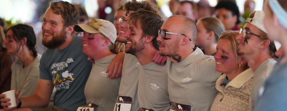 Group of smiling people with race bibs and black face paint sit closely together, arms linked. Indoor event setting with casual attire.