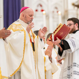 Clergy in white and gold robes during a religious service, one holding a red book. Ornate background, solemn mood in a church setting.