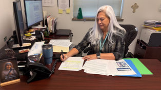 Woman with long white hair working at a desk, writing on documents. Office setting with computer, books, and a St. Teresa of Calcutta plaque.