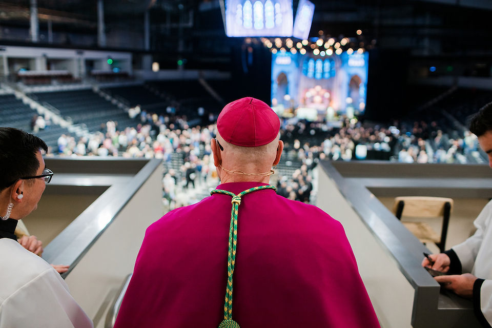 A person in a magenta robe overlooks a large gathering in an auditorium. Bright lights and a stage are visible in the background.